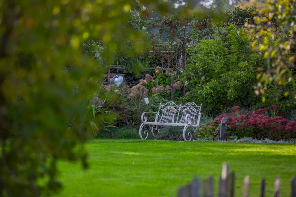 Eine elegante weiße Gartenbank steht in einem grünen, gepflegten Garten, umgeben von üppigem Pflanzenbewuchs und bunten Blumen. Im Hintergrund sind Sträucher und eine dekorative Rankhilfe zu sehen, die eine einladende und ruhige Atmosphäre schaffen.