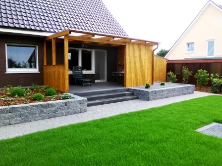 Moderne Terrasse mit Holzüberdachung an einem Einfamilienhaus, umgeben von gepflegtem Rasen und Steinbeeten. Die Terrasse ist durch eine Treppe zugänglich und bietet Platz für Gartenmöbel. Im Hintergrund sind weitere Wohnhäuser sichtbar.