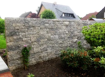 Natursteinmauer in einem Garten, umgeben von grünen Sträuchern und Blumen. Die Mauer ist aus grauen Steinen errichtet und hat eine leicht geneigte Form. Im Hintergrund sind Wohnhäuser und ein gepflegter Garten sichtbar.