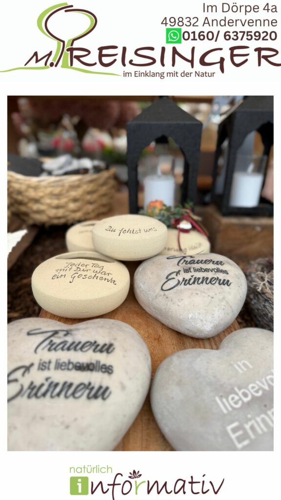 Eine Auswahl von Steinen mit verschiedenen eingravierten emotionalen Botschaften, darunter "Du fehlst uns" und "Trauer ist liebevolles Erinnern", auf einem Tisch mit Zubehör für die Dekoration und einer Laterne im Hintergrund.