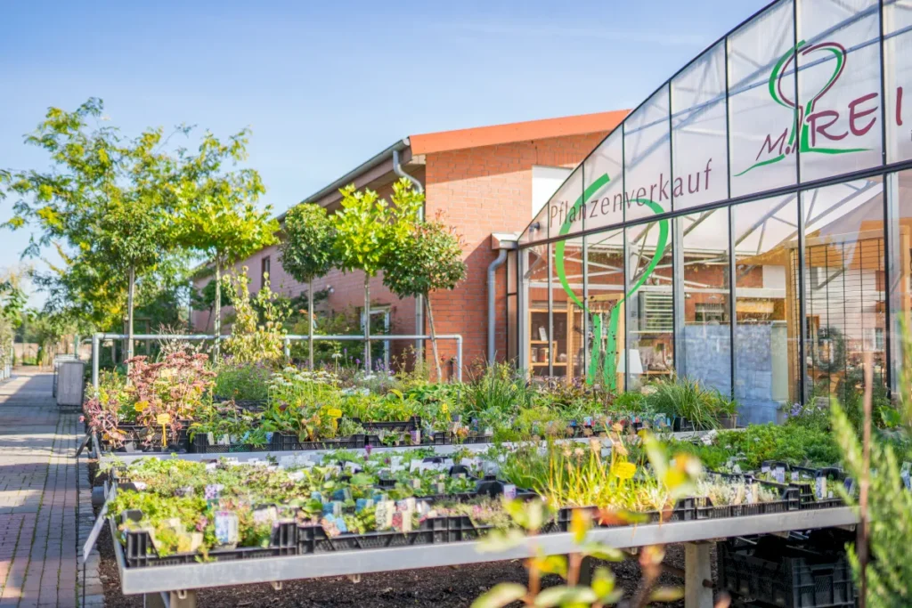 Ein Pflanzenverkauf mit bunten Pflanzen und Bäumen vor einem modernen Gebäude unter klarem Himmel.