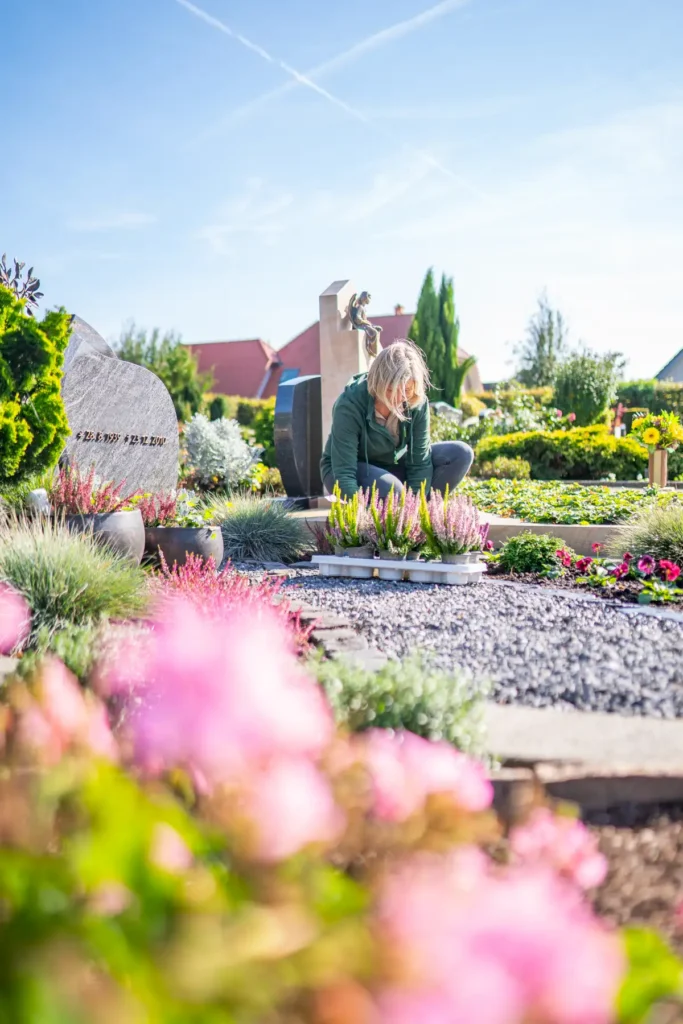 Eine Frau pflegt Blumen auf einem liebevoll gestalteten Friedhof, umgeben von bunten Pflanzen und grabsteinähnlichen Monumenten.