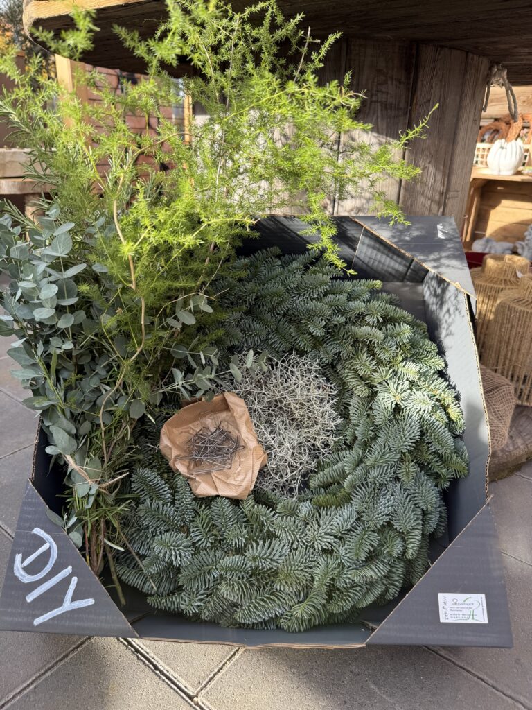 Eine DIY-Packung mit verschiedenen Pflanzenmaterialien, darunter Eukalyptus, Moos und immergrüne Zweige, in einer schwarzen Schachtel. In der Mitte befindet sich ein Papiertütchen mit Stiften zum Befestigen der Materialien, während im Hintergrund Holz- und Bastelobjekte sichtbar sind.