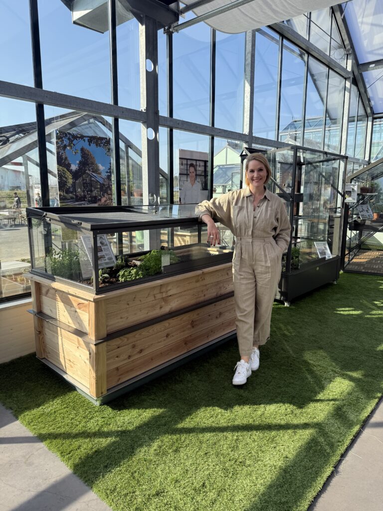 Eine Person steht in einem Gewächshaus neben einem mit Pflanzen gefüllten Holzregal. Die Person trägt einen beigen Jumpsuit und weiße Sneakers, und die Umgebung ist von viel natürlichem Licht durch große Fenster geprägt.