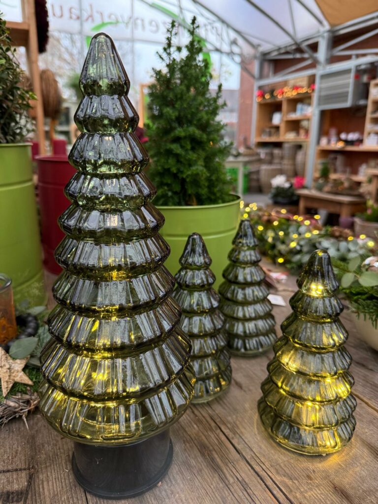 Vier gläserne Weihnachtsbäume in unterschiedlichen Größen, mit integrierten Lichtern, stehen auf einem Holztisch, umgeben von dekorativem Grün und Weihnachtsdekorationen in einem Verkaufsraum.