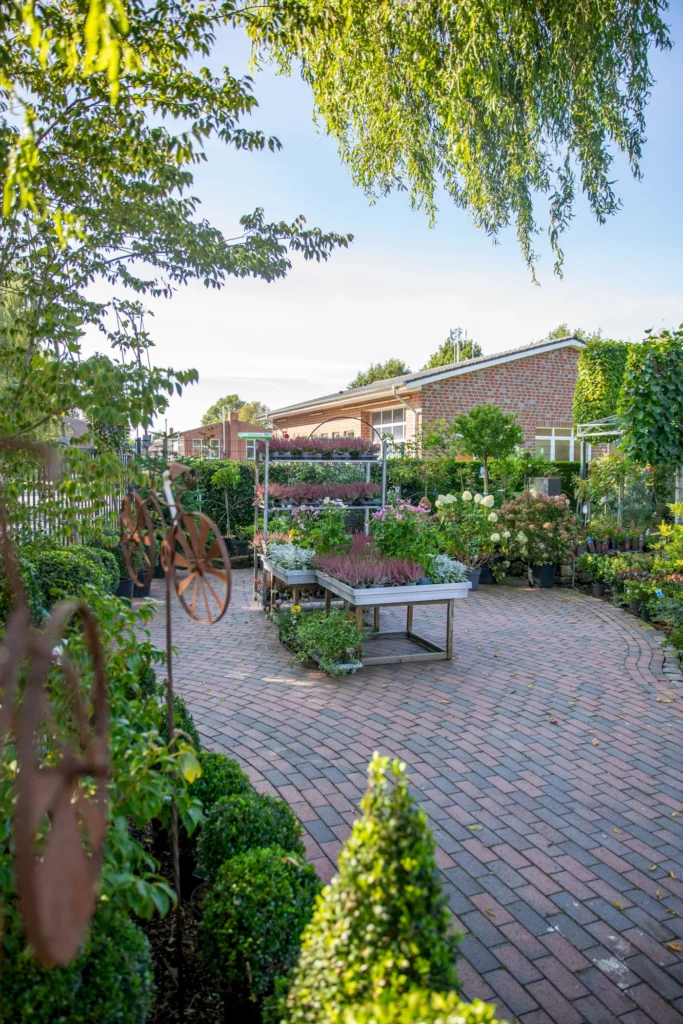 Blühender Garten mit bunten Pflanzen und Kräutern auf einem gepflasterten Weg, umgeben von grünen Hecken. Im Hintergrund ist ein Backsteingebäude und eine gemütliche Atmosphäre zu erkennen.
