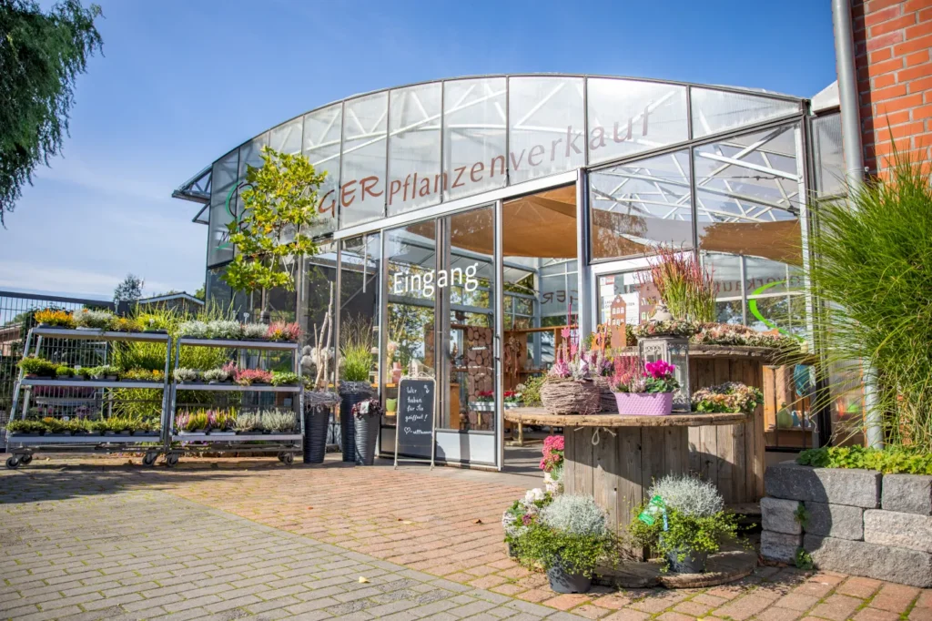 Eingang zu einem Pflanzenverkauf mit gläserner Fassade, präsentiert eine Vielzahl von Pflanzen und Blumen in dekorativen Töpfen und Regalen. Die Gestaltung umfasst Holzständer und eine einladende Atmosphäre, um Kunden anzuziehen.