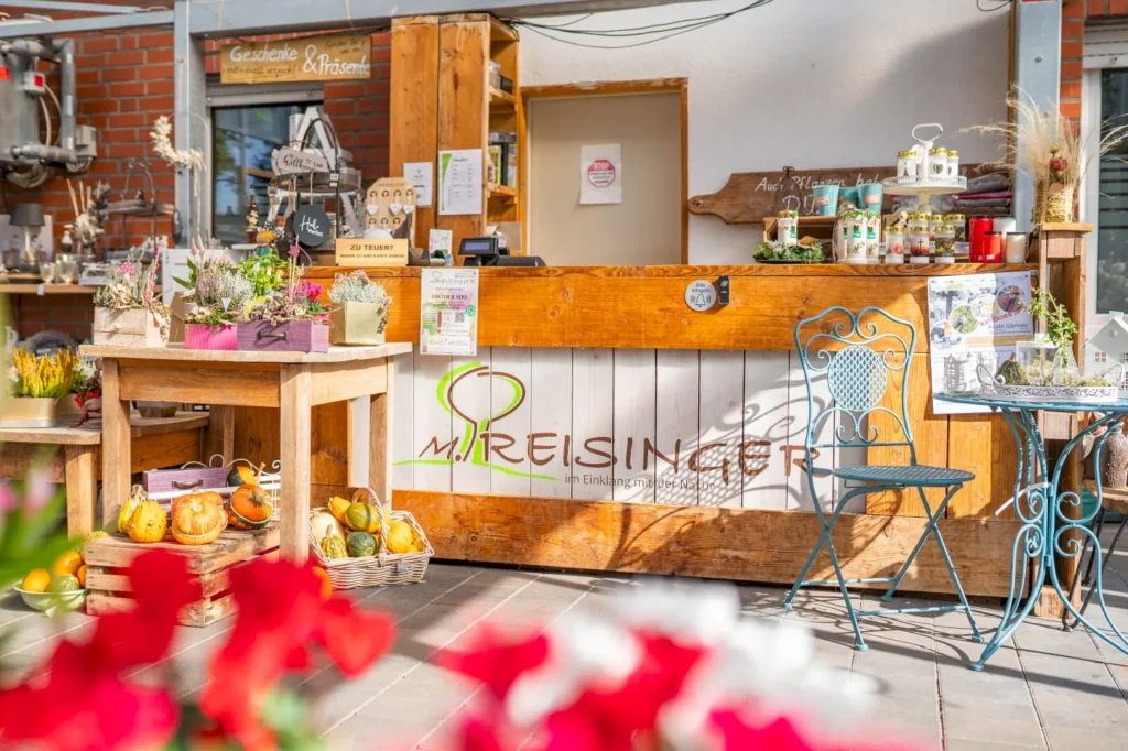 Eine einladende Verkaufsstelle mit Holztheke, bunten Pflanzen in Töpfen und frischen Gemüseprodukten. Im Hintergrund sind Geschenke und Produkte aus der Region sichtbar. Ein blauer, geschwungener Stuhl und ein Tisch ergänzen die freundliche Atmosphäre.