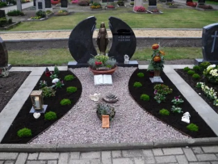 Ein liebevoll gestaltetes Grab mit einer Statue, Blumen, Landschaftsgestaltung und zwei Steinplatten im Hintergrund.