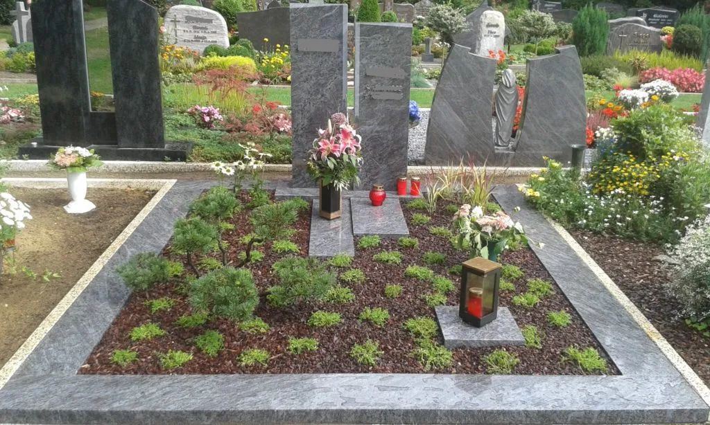 Das Bild zeigt eine liebevoll gestaltete Grabstätte, die mit verschiedenen Elementen der Garten- und Landschaftsgestaltung harmonisch in Szene gesetzt ist. Der Grabbereich ist mit dunklem Granit umrandet, was ihm eine elegante und würdige Optik verleiht. In der Mitte des Grabs sind verschiedene Steine platziert, die als Grundlage für die Grabmalgestaltung dienen.
Um die Steine herum sind sorgfältig ausgewählte Pflanzen angeordnet, die dem Grab Leben