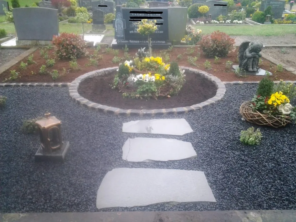 Gepflegter Friedhofsbereich mit einem runden Blumenbeet, umgeben von bunten Blumen und Sträuchern. Im Vordergrund führen graue Steinplatten über einen mit Kies bedeckten Weg zu einer Laterne. Eine Statue eines Engels steht neben dem Blumenbeet, während im Hintergrund weitere Grabsteine sichtbar sind.