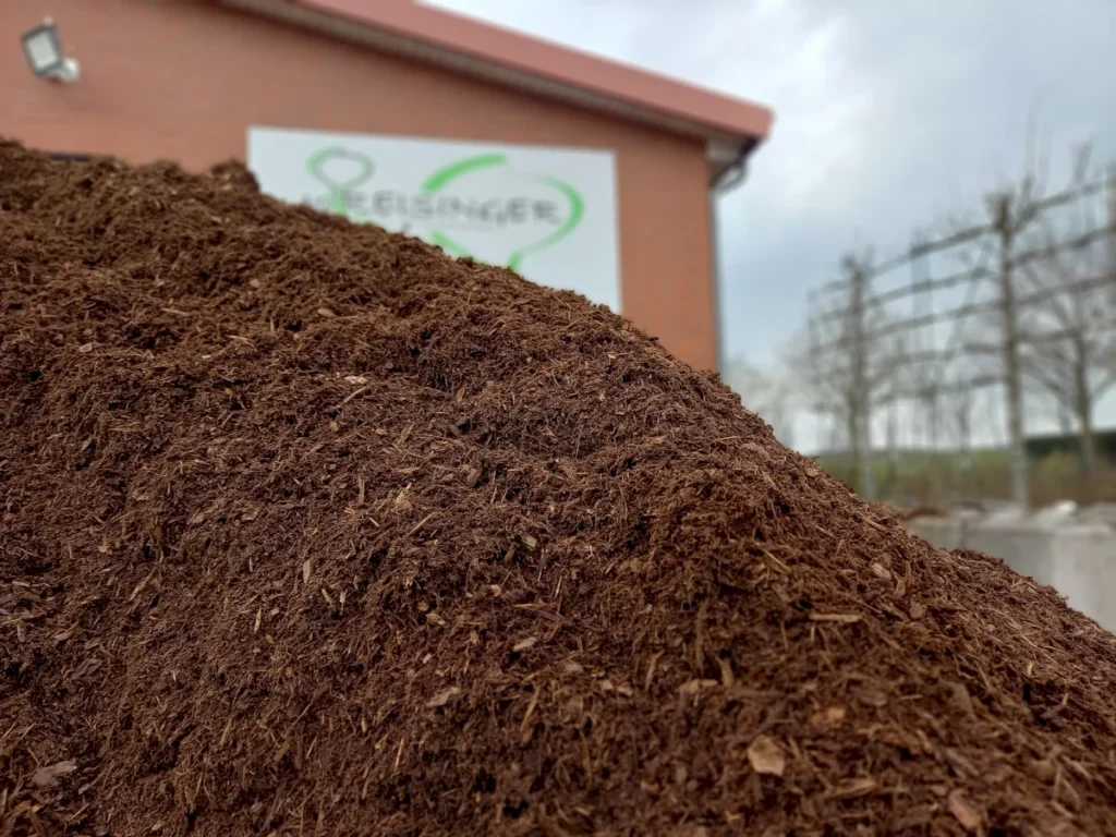 Dunkle, frische Erde liegt in einer kleinen Hügelform im Vordergrund, während im Hintergrund ein Gebäude mit einem grünen Logo sichtbar ist. Der Himmel ist bewölkt, was auf bevorstehendes Wetter hinweist.